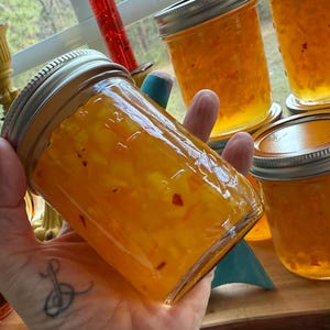 May include: A hand holding a clear glass jar filled with orange-colored jam. The jam has small red flecks throughout. Several other jars of jam are visible in the background, all with silver lids. The jars are on a wooden surface near a window.