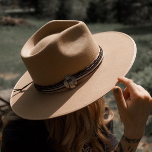May include: A tan felt fedora hat with a brown leather band and a silver bird design. The hat is being held by a person with tattoos on their arms.