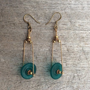 Puede incluir: Un par de pendientes de oro con círculos de vidrio azul verdoso que cuelgan de la parte inferior. Los pendientes están hechos de alambre y tienen pequeñas cuentas doradas en la parte superior.