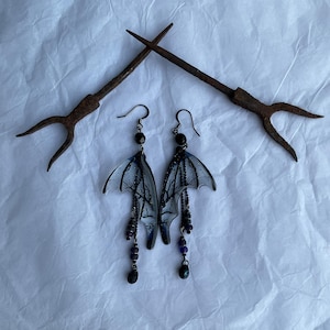 May include: A pair of gothic-style earrings. The earrings have translucent, wing-shaped pendants with black tree branch designs and dangling beads in black and purple. The earrings are suspended from black hooks.