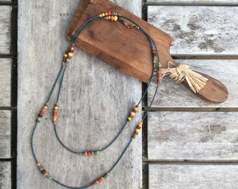 May include: A double strand necklace with colourful beads in shades of brown, yellow, orange, green, and blue. The necklace is made of dark blue cord and is lying on a wooden surface.