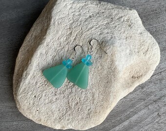 May include: A pair of teal green sea glass earrings with silver hooks and teal blue beads. The earrings are displayed on a light grey rock.