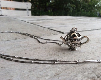 May include: A silver necklace with a teapot pendant. The teapot is detailed with a filigree design and has a small spout and handle. The necklace is on a wooden surface.