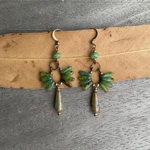 May include: A pair of handmade earrings with a unique design. The earrings feature a gold-colored hook, a green bead, and a fan-shaped cluster of green glass beads. The earrings also have a teardrop-shaped bead at the bottom. The earrings are displayed on a leaf.