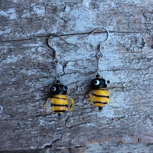May include: A pair of bee earrings. Each earring has a black head with white eyes and a yellow and clear striped body. The earrings hang from silver-coloured hooks.