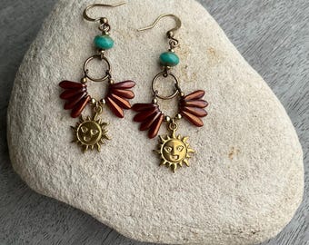 May include: Handmade earrings with a sun motif. The earrings have a gold finish, a turquoise bead, a fan-shaped element, and a smiling sun pendant. The earrings are presented on a light-coloured stone. The earrings are a unique piece of jewellery.