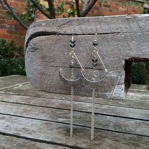 Op de afbeelding: Een paar zilveren oorbellen met een halve maan ontwerp en een hangende ketting. De oorbellen hebben een kleine groene steen en een geometrisch patroon.