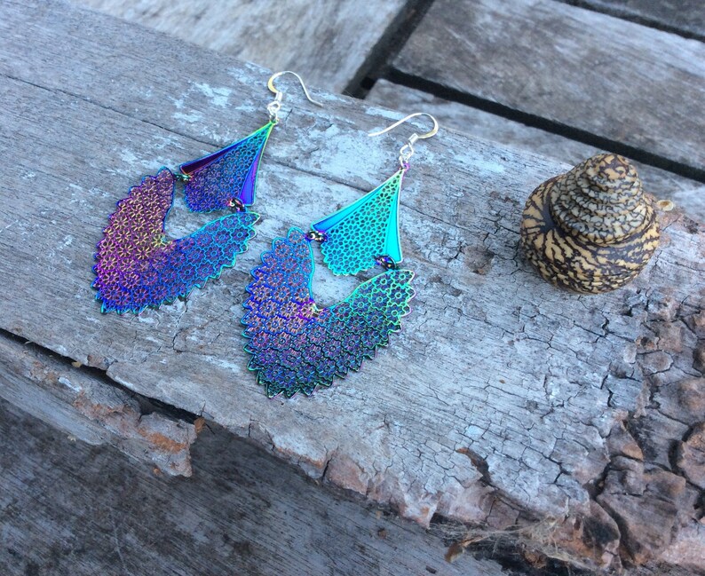 May include: A pair of iridescent earrings with a delicate, intricate design. The earrings are made of metal and have a silver tone. They are shaped like a teardrop and have a layered design.