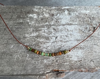 Può includere: Una collana fatta a mano con un cordino marrone e una serie di piccole perline colorate. Le perline includono sfumature di verde, marrone, arancione e oro, creando un'estetica naturale e terrosa. La collana è esposta su una superficie di legno invecchiato.