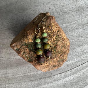 May include: A pair of dangle earrings with green, brown, and burgundy beads. The earrings have a brass finish and are hanging from a rock.