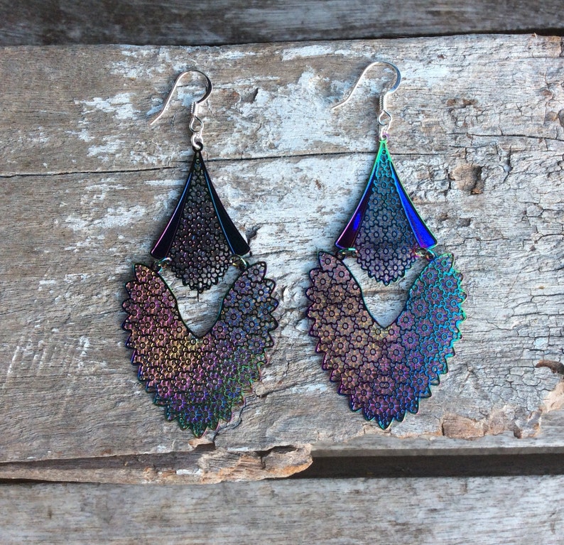 May include: A pair of iridescent earrings with a layered design. The earrings are made of metal and have a geometric shape. The earrings are a dark purple, blue, and green color.