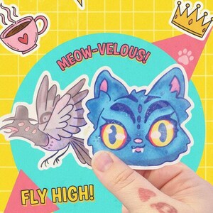 Peut inclure: Une main tenant un autocollant avec une illustration de chat bleu et le texte "MEOW-VELOUS!". D'autres autocollants incluent un oiseau, une couronne et le texte "CREATIVE VIBES!" et "STICKER FUN!" sur un fond jaune quadrillé.
