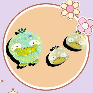 May include: Three green and yellow duck-shaped resin pins with glitter accents. The largest duck has a crown on its head.