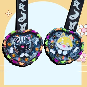 May include: Two heart-shaped pendants with cartoon cat and dog designs. The black cat pendant has a black background with a colorful border. The dog pendant has a yellow and white dog design with a colorful border. Both pendants have black ribbons with white ghost designs.