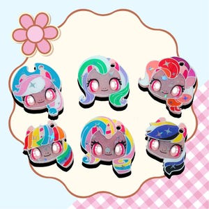May include: Six colorful, glittery cartoon character charms with large eyes and various hair colors, including pink, blue, green, and rainbow. Each charm has a small hole at the top for attaching to a keychain or other accessory. The background is a light blue and pink checkered pattern.
