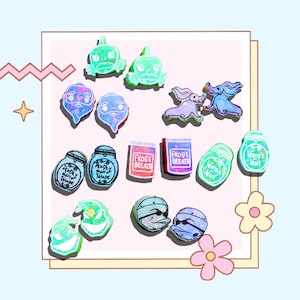 Può includere: Una collezione di 14 ciondoli in resina colorati con vari personaggi e design, tra cui un fantasma, una rana, un barattolo e un teschio. I ciondoli sono iridescenti e hanno una finitura scintillante.