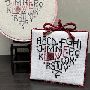 May include: Two embroidery hoops with a white fabric background and black stitching. The stitching forms a heart shape with the letters of the alphabet inside. The word "LOVE" is stitched in red in the centre of the heart. The smaller hoop is framed with a burgundy ribbon.