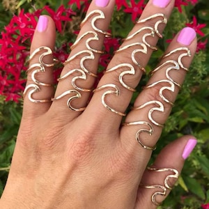 May include: A hand wearing multiple gold-colored wave-shaped rings. The rings are on the fingers, with a pink nail polish. The background features red flowers and green foliage.