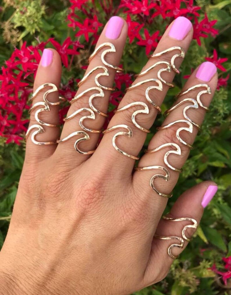 May include: A hand wearing multiple gold-colored wave-shaped rings. The rings are on the fingers, with a pink nail polish. The background features red flowers and green foliage.