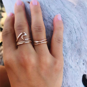 May include: A hand wearing several gold rings. One ring is shaped like a wave, another is a twisted rope design, and the third is a criss-cross design. The rings are on a hand with light pink nail polish.