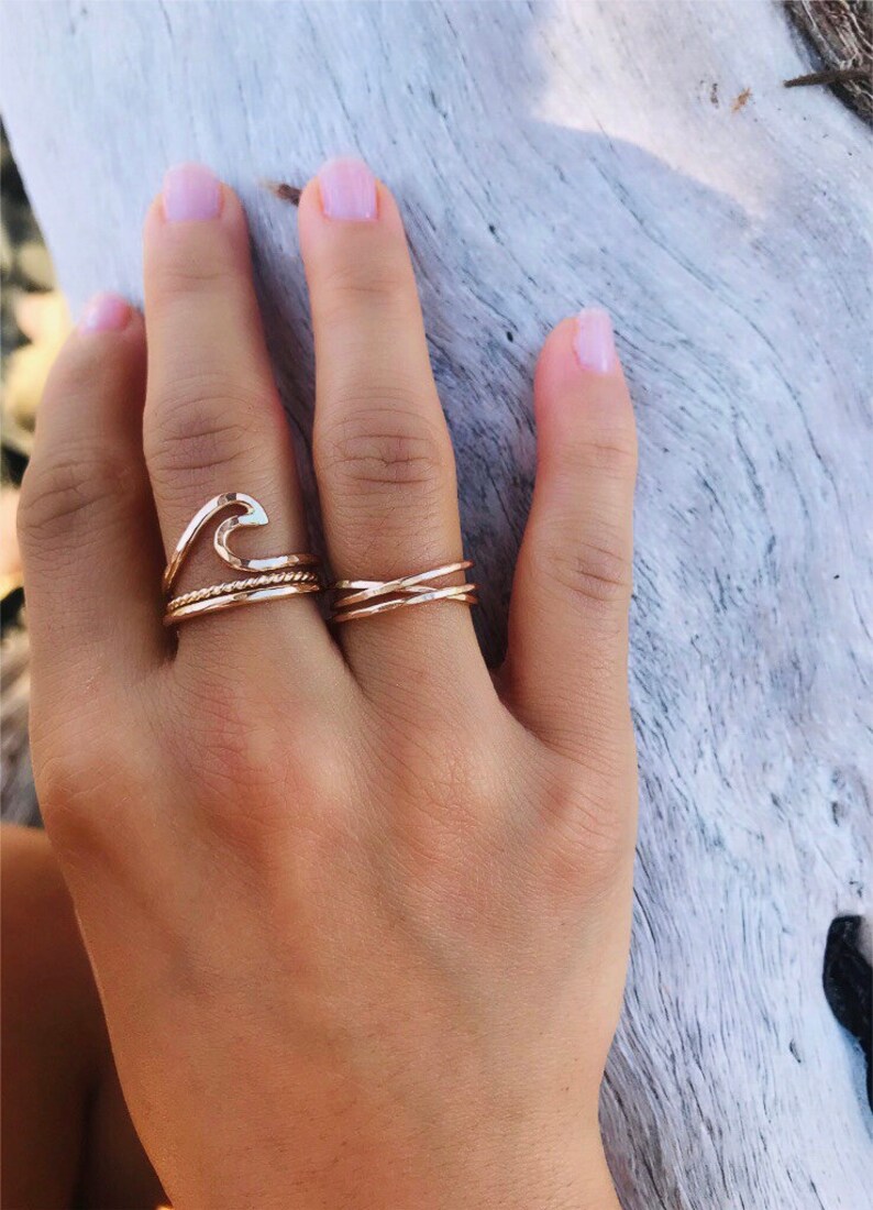May include: A hand wearing several gold rings. One ring is shaped like a wave, another is a twisted rope design, and the third is a criss-cross design. The rings are on a hand with light pink nail polish.