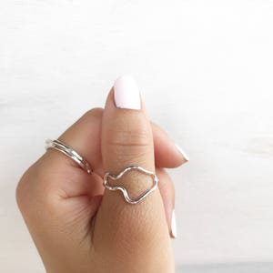 May include: A silver ring with a wavy, abstract design. The ring is worn on a person's thumb.