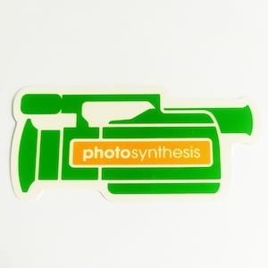 May include: A green and white sticker in the shape of a video camera with the word "photosynthesis" in orange and white. The sticker has a white background and a green outline.