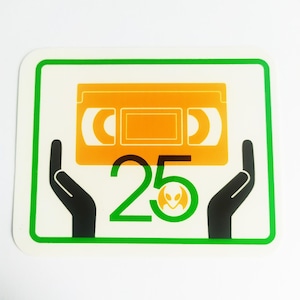 May include: A sticker with a green border features a stylized orange VHS tape graphic. Below the tape is the number 25 in green, with a small alien head logo. Black hands appear to be holding the tape.