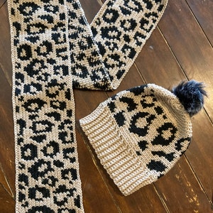 May include: A crocheted hat and scarf set in a beige and black leopard print pattern. The hat has a fluffy pom pom on top.