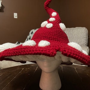 Puede incluir: Sombrero de hongo de ganchillo hecho a mano, rojo con lunares blancos y un borde blanco. El sombrero se exhibe en una cabeza de maniquí. El tallo del sombrero es de color beige claro. Un accesorio caprichoso.