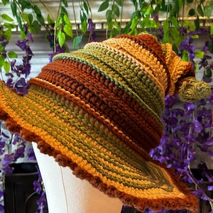 Puede incluir: Un sombrero de ganchillo con una corona puntiaguda y un ala ancha y ondulada. El sombrero está construido con bandas horizontales de hilo en tonos alternos de amarillo mostaza, verde bosque y marrón oscuro, con un borde texturizado y festoneado.