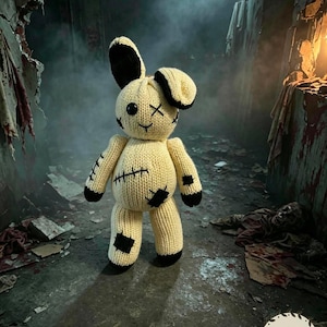 Puede incluir: Un conejo de peluche tejido de color crema con puntas de orejas negras, patas negras y parches negros cosidos en el cuerpo y las extremidades. La cara tiene un ojo de botón negro, un ojo en forma de X y una sonrisa simple bordada.