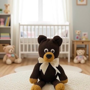 Pode incluir: Um urso de pelúcia castanho feito à mão com um laço creme e detalhes amarelos. O urso tem olhos grandes e arregalados e está sentado em um tapete creme. O urso tem aproximadamente 30 cm de altura.