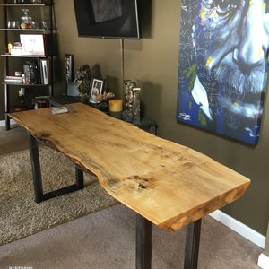 May include: A live edge wood table with a natural wood finish and black metal legs. The table is in a room with a television, a bookshelf, and a painting of Albert Einstein with mathematical equations.