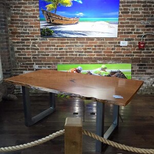 May include: A large, live edge wooden table with a natural wood finish and metal legs. The table is displayed in a room with brick walls and a rope railing. The table is made by Kentucky Live Edge.
