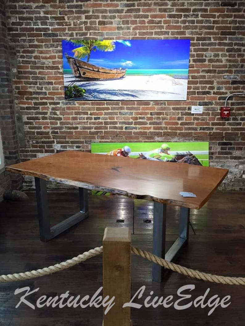 May include: A large, live edge wooden table with a natural wood finish and metal legs. The table is displayed in a room with brick walls and a rope railing. The table is made by Kentucky Live Edge.