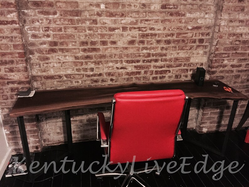May include: A dark wood live edge desk with black metal legs and a red office chair. The desk is in front of a brick wall.