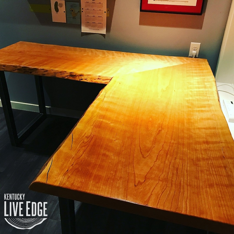 May include: A large, live edge, L-shaped desk made of light wood with a natural, unfinished finish. The desk has a black metal frame and is illuminated by a warm light.