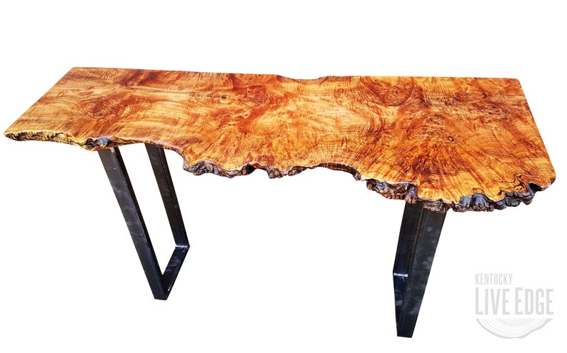 May include: A live edge wood console table with a natural wood finish and a unique, irregular shape. The table has a black metal frame with two legs on each side. The table is made by Kentucky Live Edge.
