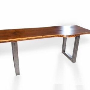 May include: A long, rectangular wooden table with a live edge top and a natural wood finish. The table has two metal legs with a square design.