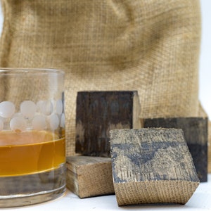 May include: A glass of amber-colored liquid sits on a white surface in front of four wooden blocks. The blocks are various shades of brown and have a rough, textured surface. A burlap sack sits behind the blocks.