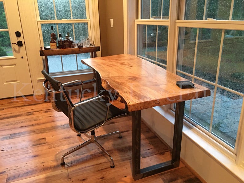 May include: A live edge wood desk with a natural wood finish and a metal frame. The desk is positioned in front of a window with a view of trees. A black office chair is positioned next to the desk.