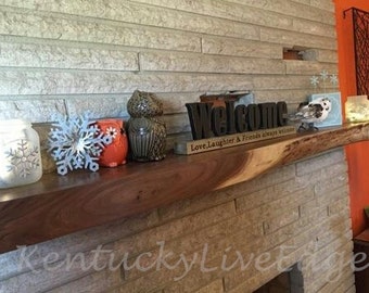 May include: A wooden mantelpiece with a variety of decorative items, including a welcome sign, two ceramic owls, a snowflake ornament, and a jar with a snowflake design. The sign reads "Welcome, Love, Laughter & Friends always welcome".