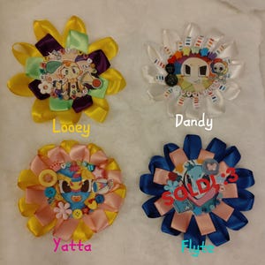 May include: Four colorful, flower-shaped ribbon brooches. Each brooch features a central cartoon character design with text labels: "Looney", "Dandy", "Yatta", and "Flyte". The "Flyte" brooch has "SOLD" text.