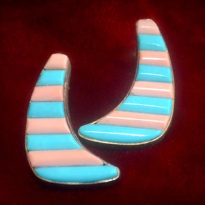 May include: A pair of crescent-shaped earrings with alternating horizontal stripes of light blue and pale pink. The earrings have a gold-colored trim and are set against a deep red background.
