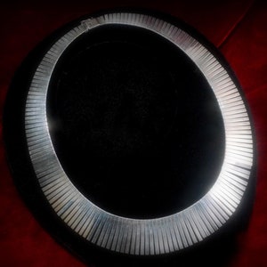 May include: A silver-coloured collar necklace with a ribbed design. The necklace is circular and lies on a black surface, set against a red background. The jewellery piece is a statement accessory.