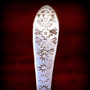 May include: A silver spoon with a decorative handle featuring a geometric pattern of stars and triangles. The handle is shaped like a teardrop.