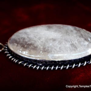 May include: A silver pendant with a large, oval-shaped, clear quartz stone set in a silver bezel. The stone has a cloudy, milky appearance. The pendant is on a red velvet background.