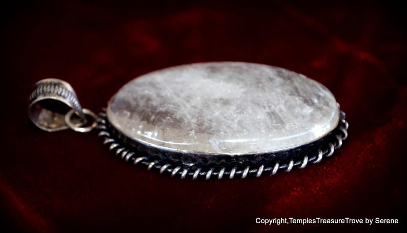 May include: A silver pendant with a large, oval-shaped, clear quartz stone set in a silver bezel. The stone has a cloudy, milky appearance. The pendant is on a red velvet background.