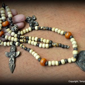 Puede incluir: Un collar de cuentas blancas y negras con cuentas naranjas, un colgante de ángel de bronce, un colgante de rana de bronce y un colgante de placa de bronce.